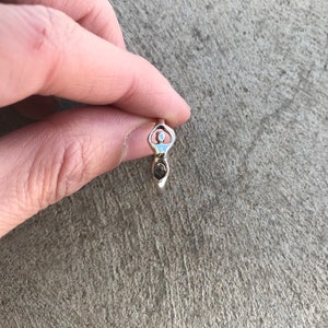 Peut inclure: Une bague en argent repr&eacute;sentant une d&eacute;esse stylis&eacute;e. La bague pr&eacute;sente une forme f&eacute;minine centrale avec les bras lev&eacute;s et un d&eacute;tail en spirale &agrave; la base. La bague est tenue sur une surface grise textur&eacute;e.