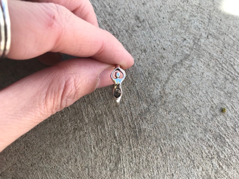 Peut inclure: Une bague en argent repr&eacute;sentant une d&eacute;esse stylis&eacute;e. La bague pr&eacute;sente une forme f&eacute;minine centrale avec les bras lev&eacute;s et un d&eacute;tail en spirale &agrave; la base. La bague est tenue sur une surface grise textur&eacute;e.