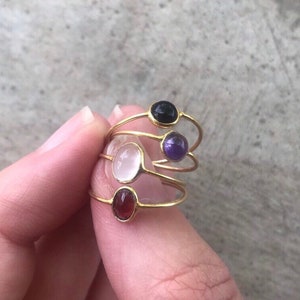 May include: Four gold rings with different colored gemstones. The rings are stacked on top of each other. The gemstones are a white oval, a red oval, a purple oval, and a black oval.