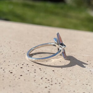 May include: A silver-colored ring with a star-shaped design. The star has a black center and pink and silver-colored points. The ring is set against a neutral background with a blurred green backdrop.