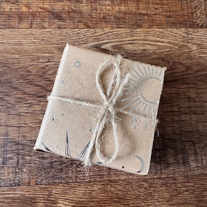 Peut inclure: Un paquet carr&eacute; envelopp&eacute; dans du papier brun, attach&eacute; avec de la ficelle. Le papier est d&eacute;cor&eacute; de motifs c&eacute;lestes argent&eacute;s, dont un soleil, une lune et des &eacute;toiles. Le paquet repose sur une surface en bois.