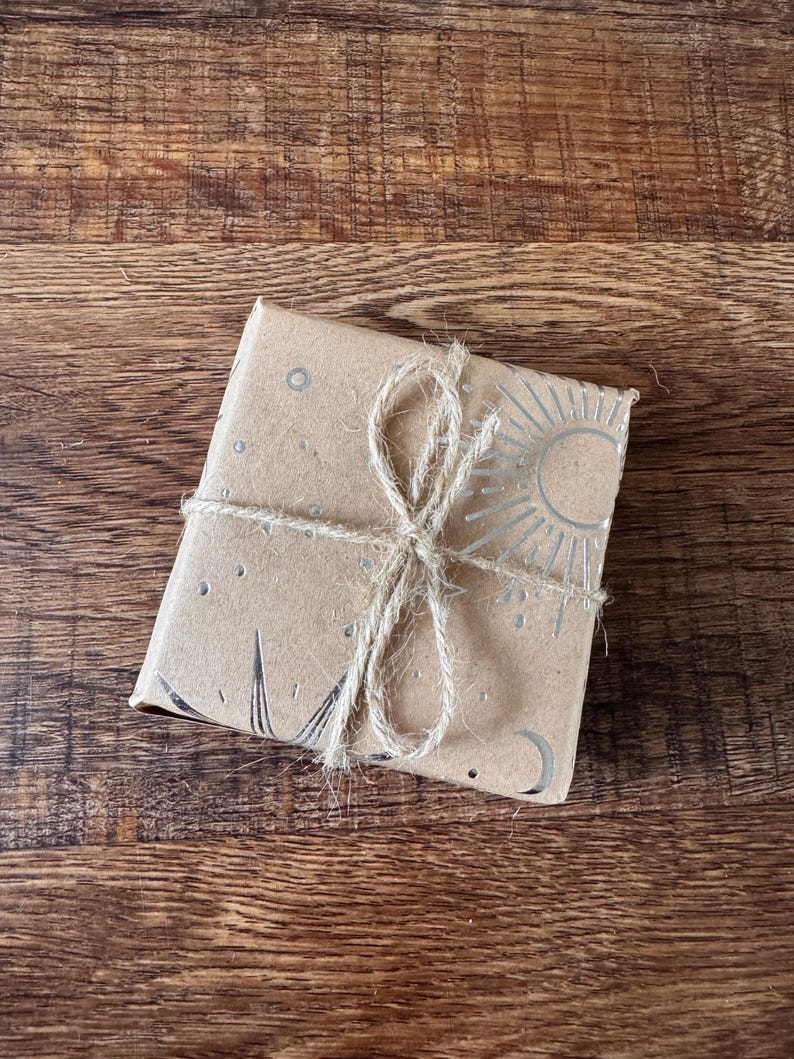 Peut inclure: Un paquet carr&eacute; envelopp&eacute; dans du papier brun, attach&eacute; avec de la ficelle. Le papier est d&eacute;cor&eacute; de motifs c&eacute;lestes argent&eacute;s, dont un soleil, une lune et des &eacute;toiles. Le paquet repose sur une surface en bois.