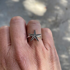 May include: A silver ring featuring a starfish design. The starfish has a textured surface and is set on a thin band. The ring is worn on a finger, showcasing the jewelry's details and design.