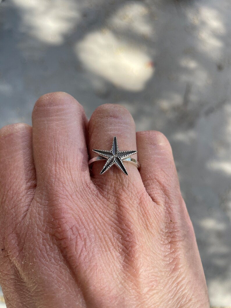 May include: A silver ring featuring a starfish design. The starfish has a textured surface and is set on a thin band. The ring is worn on a finger, showcasing the jewelry's details and design.