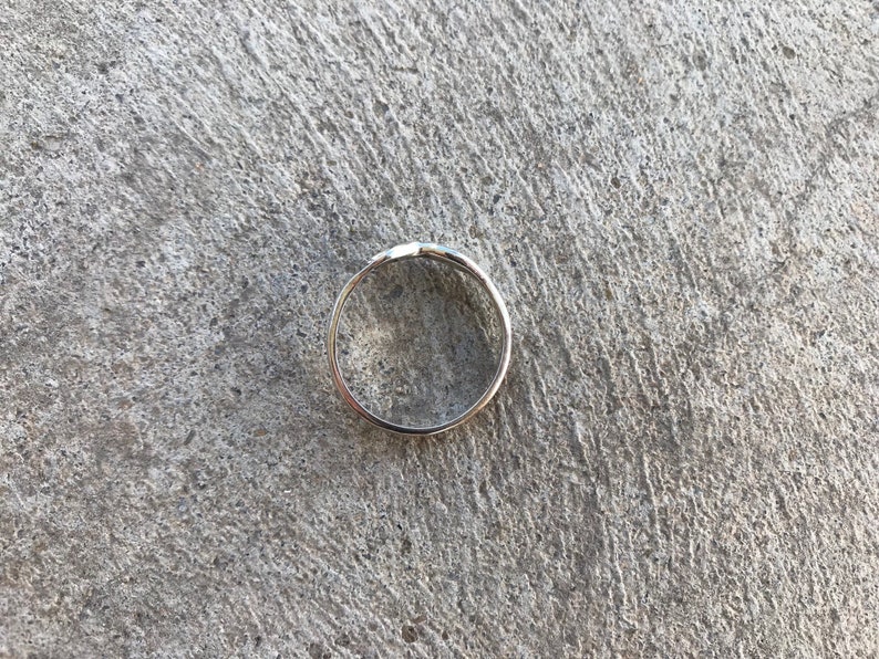 Peut inclure: Une bague en argent au design simple et &eacute;l&eacute;gant. La bague est circulaire avec un petit d&eacute;tail en relief sur le dessus. La bague est pos&eacute;e sur une surface en b&eacute;ton grise textur&eacute;e.