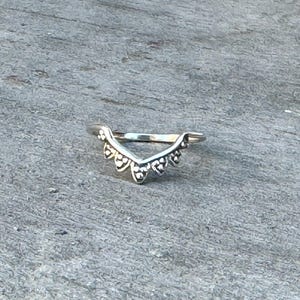 Pode incluir: Um anel de prata com um design delicado em forma de V. O anel é adornado com pequenos acentos redondos, espaçados uniformemente, ao longo das bordas.