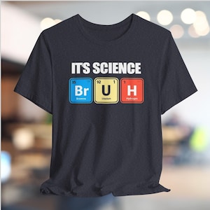 May include: Dark grey t-shirt featuring the text "IT'S SCIENCE" above the periodic table elements Br, U, and H. The elements are in blue, yellow, and red boxes. The shirt is made of a soft material.