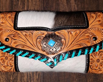 May include: A rectangular wallet featuring a combination of white and brown cowhide leather and tan tooled leather with floral patterns. The wallet is accented with bright turquoise lacing along the edges and a central turquoise stone concho.