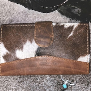 May include: A rectangular wallet featuring a brown and white cowhide pattern on the upper section and a solid brown leather lower panel. The wallet includes a central brown leather snap closure tab and visible stitching along the edges.