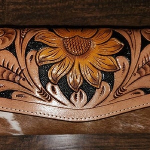 May include: A tan leather wallet with a sunflower design. The sunflower is embossed and colored in shades of yellow and brown. The wallet has a cowhide bottom and a flap closure.
