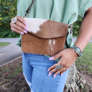 May include: A brown and white cowhide crossbody bag with a flap closure. The bag is worn over a light green shirt and blue jeans.