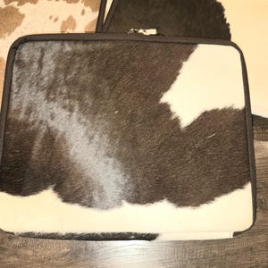 May include: Laptop sleeve with a cowhide pattern in black, white, and brown. The sleeve has a black border and a zipper. The cowhide pattern is visible on the front of the sleeve. Other sleeves with similar patterns are visible in the background.