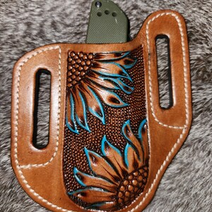 Puede incluir: Una funda de cuchillo de cuero marr&oacute;n con un lazo para el cintur&oacute;n y un dise&ntilde;o de girasol tallado en marr&oacute;n y turquesa.