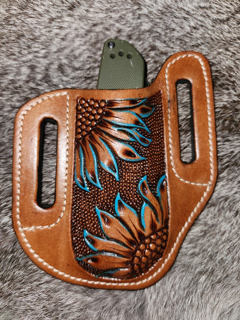 Puede incluir: Una funda de cuchillo de cuero marr&oacute;n con un lazo para el cintur&oacute;n y un dise&ntilde;o de girasol tallado en marr&oacute;n y turquesa.