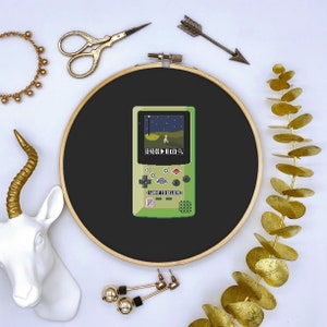 May include: A green and black pixelated Game Boy-style handheld game console with a screen displaying a pixelated image of a UFO and the text "I WANT TO BELIEVE".