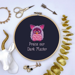 May include: Embroidered hoop art featuring a pink and purple pixelated Furby with the text "Praise our Dark Master" on a dark blue background.