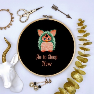 Puede incluir: Un aro de bordado negro con una imagen pixelada rosa y verde de un juguete Furby. El texto "Go to Sleep Now" está escrito en rosa debajo de la imagen.