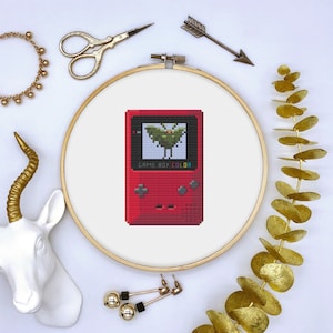 May include: A red Game Boy Color with a green pixelated creature on the screen. The Game Boy Color is in a wooden embroidery hoop.