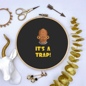 May include: A cross-stitch embroidery hoop with a black background features a pixelated image of a character and the text "IT'S A TRAP!" in yellow. The hoop is surrounded by gold accessories, including scissors, an arrow, and a bracelet.