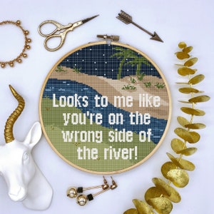 Puede incluir: Un aro de bordado redondo, verde y marrón con una escena de río azul y arena. El texto "Looks to me like you're on the wrong side of the river!" está bordado en hilo blanco.