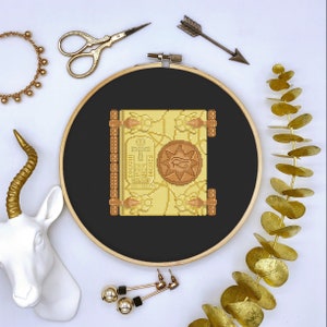 May include: A cross-stitch embroidery hoop with a black fabric background and a yellow and brown embroidered image of an ancient Egyptian book with hieroglyphics and an ankh symbol.