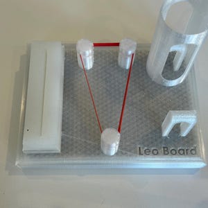 May include: A 3D-printed clear plastic model with a rectangular base. It features white cylindrical structures connected by a red string, a tall cylinder, a rectangular piece, and the text "Leo Board".