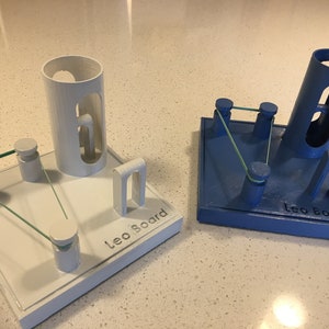 Puede incluir: Dos Leo Boards impresos en 3D, uno blanco y otro azul, se muestran uno al lado del otro. Cada tablero tiene una base cuadrada con postes cilíndricos y un cilindro alto con lados abiertos. Hay gomas elásticas verdes.