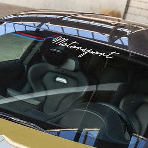 Puede incluir: Primer plano del parabrisas de un coche con una calcomanía blanca de "Motorsport" en la parte superior. El parabrisas está tintado y refleja el interior del coche, que tiene asientos de cuero negro.