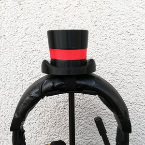 Può includere: Supporto per cuffie nero con un cappello a cilindro nero e una fascia rossa. Il supporto è in plastica e ha un'asta di metallo.