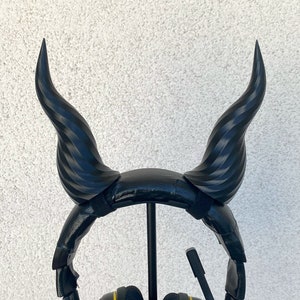 Puede incluir: Auriculares negros con detalles amarillos en los auriculares, unidos a una diadema negra con dos cuernos negros impresos en 3D. Los auriculares están en un soporte negro.