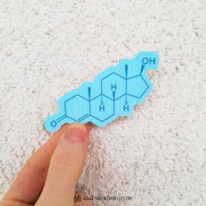 May include: A light blue sticker featuring a chemical structure diagram. The sticker has a textured surface and the chemical structure is printed in a darker shade of blue. The sticker is held by a hand.