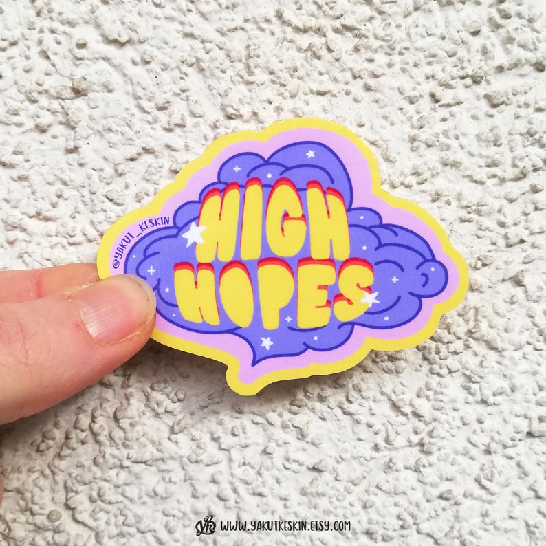 Puede incluir: Una pegatina amarilla y morada con el texto "HIGH HOPES" en una fuente retro. La pegatina tiene forma de nube con estrellas blancas.