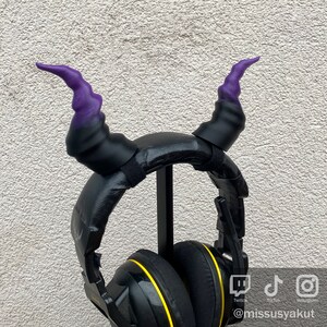 May include: Black headphones with yellow accents and purple horns attached to the headband. The headphones are on a stand.