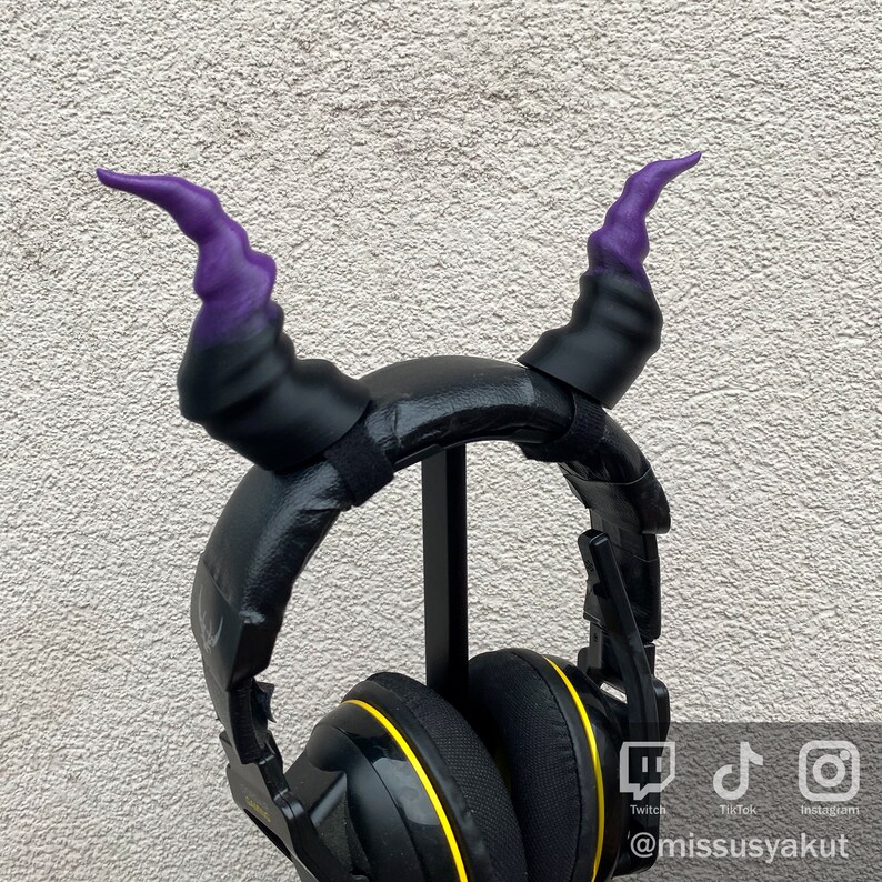May include: Black headphones with yellow accents and purple horns attached to the headband. The headphones are on a stand.