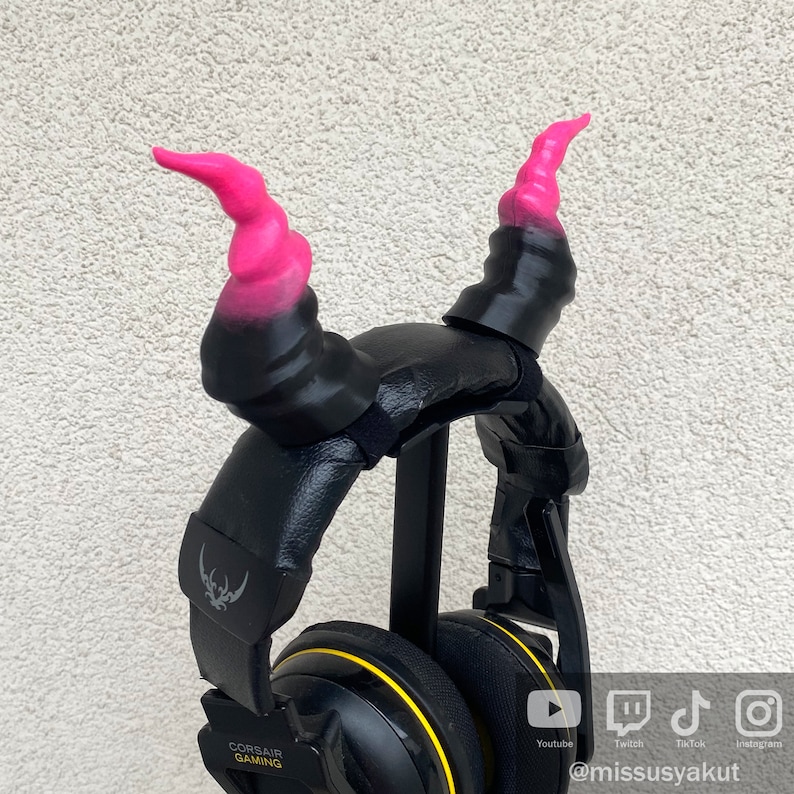 May include: Black gaming headset with pink and black devil horns attached to the headband. The headset has a yellow stripe around the earcups and the text "CORSAIR GAMING" is printed on the side.