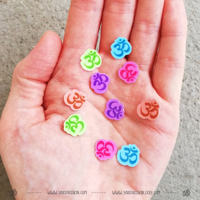 May include: A hand holding a variety of colorful stickers with the Om symbol. The stickers are in various colors, including pink, blue, green, orange, and purple.