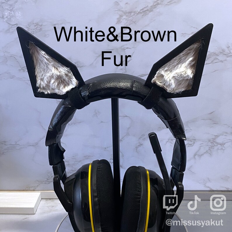 May include: Black headphones with yellow accents and white and brown fur cat ears attached to the headband. The headphones are on a stand with social media icons and the text "@missusyakut" below.