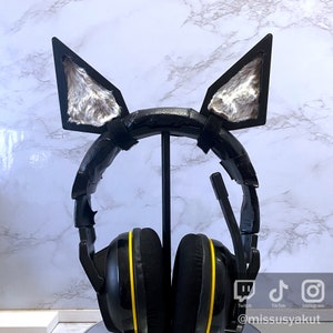 May include: Black gaming headset with yellow accents and furry gray cat ears attached to the headband. The headset is on a stand and the social media icons for Twitch, TikTok, and Instagram are visible in the lower right corner of the image.