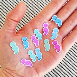 May include: A collection of colorful semicolon stickers in pink, blue, and turquoise. The stickers are arranged in a semi-circle on a person's hand.