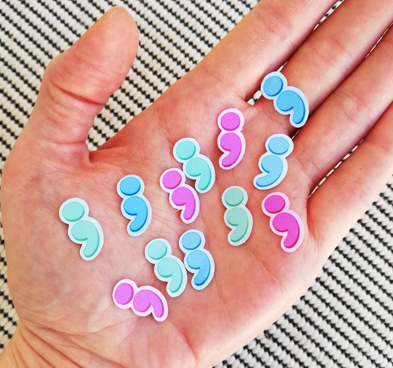 May include: A collection of colorful semicolon stickers in pink, blue, and turquoise. The stickers are arranged in a semi-circle on a person's hand.