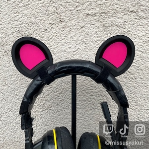 Könnte beinhalten: Schwarze Kopfhörer mit rosa Katzenohren, die am Kopfbügel befestigt sind. Die Kopfhörer stehen auf einem Ständer und haben gelbe Akzente an den Ohrmuscheln. Der Text "@missusyakut" ist unter den Kopfhörern sichtbar.