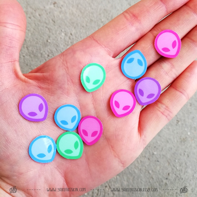 May include: A hand holding a variety of colorful alien head stickers. The stickers are in shades of pink, blue, green, and purple.
