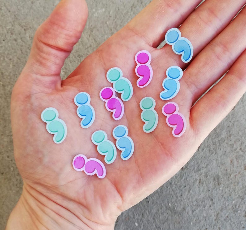 May include: A hand holding a variety of colorful semicolon stickers. The stickers are in shades of blue, pink, and green.