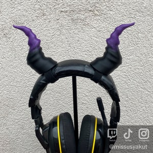 May include: Black headphones with purple and black devil horns attached to the headband. The headphones have yellow accents around the earcups. The headphones are on a stand.