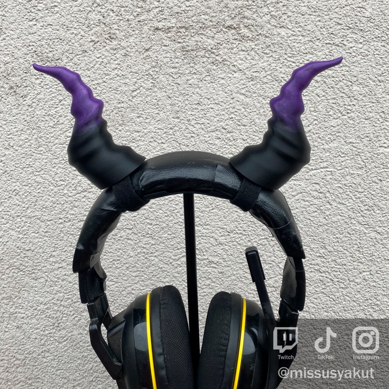 May include: Black headphones with purple and black devil horns attached to the headband. The headphones have yellow accents around the earcups. The headphones are on a stand.