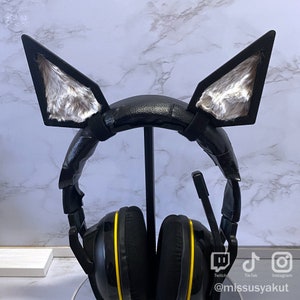 May include: Black gaming headset with yellow accents and furry cat ear attachments. The headset is on a stand and has logos for Twitch, TikTok, and Instagram.