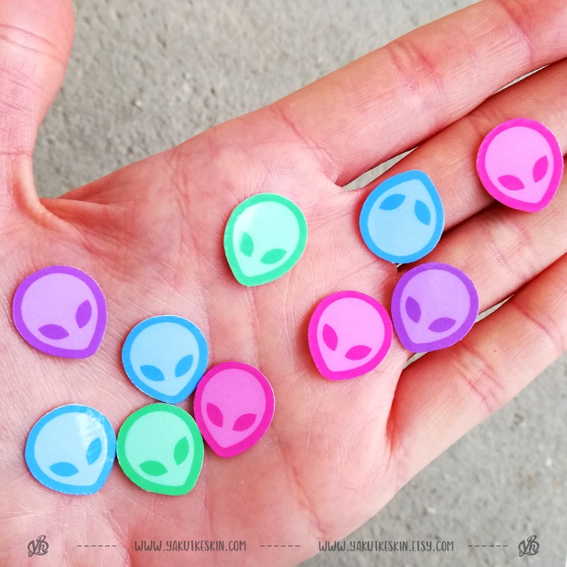 May include: A hand holding a variety of colorful alien head stickers. The stickers are in shades of pink, blue, green, and purple. The stickers are all the same size and shape, and they feature a simple alien head design.