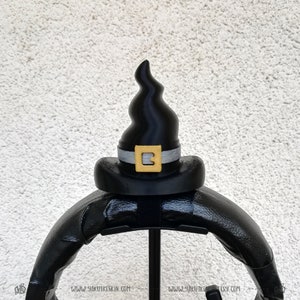 May include: Black witch hat with a gold buckle and a silver band. The hat is on a black stand.