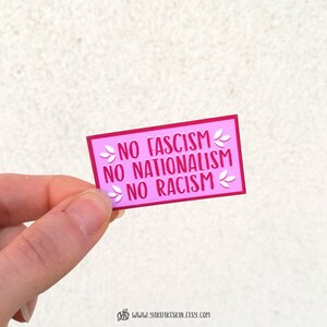 K&ouml;nnte beinhalten: Rosafarbener Aufkleber mit dem Text "NO FASCISM NO NATIONALISM NO RACISM" in roten Buchstaben mit wei&szlig;en floralen Akzenten.