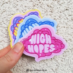 Puede incluir: Tres pegatinas coloridas con un dise&ntilde;o de nube. La pegatina superior es amarilla con una nube morada y el texto "Stay Wild". La pegatina del medio es azul con una nube azul y el texto "Dream Big". La pegatina inferior es rosa con una nube rosa y el texto "High Hopes".